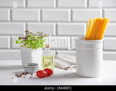 Raw spaghetti with tomatoes, basil, pepper and olive oil on a white brick background. Traditional ingredients for making Italian pasta. Front view. Stock Photo
