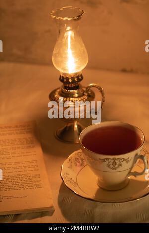 photo of a glowing oil lamp and a retro cup of tea on a saucer standing next to an old open book Stock Photo