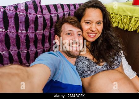 Latin couple selfie portrait, caucasian adult man with beard and blue eyes with his wife, pregnant brazilian adult woman smiling, happy sitting on sof Stock Photo