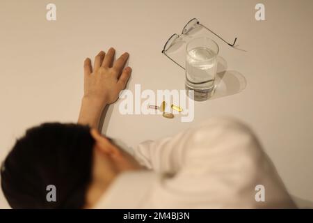 Tired woman is putting her head down on the desk, before taking medicine with cup of water on the white table, girl is taking nutritional supplement Stock Photo
