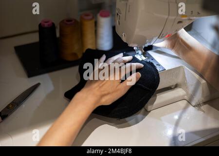 Woman is sewing black beanie hat on a sewing machine, using a sewing machine on the sewing table Stock Photo