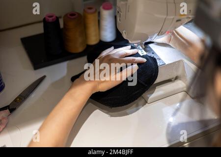 Woman is sewing black beanie hat on a sewing machine, using a sewing machine on the sewing table Stock Photo
