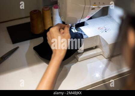 Woman is sewing black beanie hat on a sewing machine, using a sewing machine on the sewing table Stock Photo