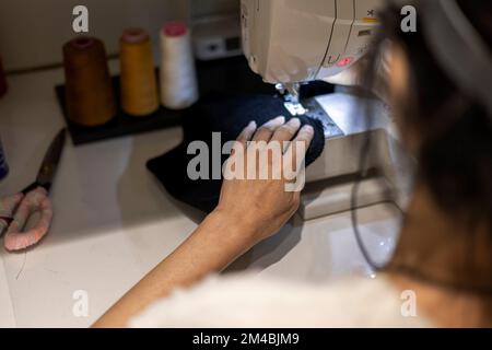 Woman is sewing black beanie hat on a sewing machine, using a sewing machine on the sewing table Stock Photo