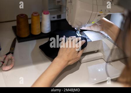 Woman is sewing black beanie hat on a sewing machine, using a sewing machine on the sewing table Stock Photo