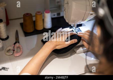 Woman is sewing black beanie hat on a sewing machine, using a sewing machine on the sewing table Stock Photo