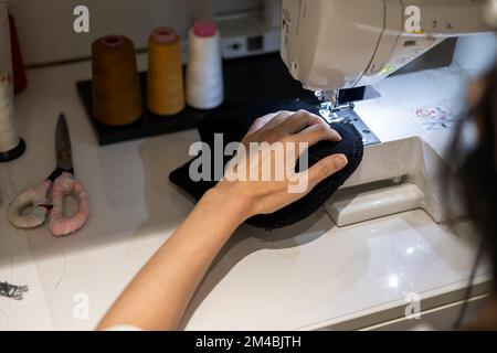 Woman is sewing black beanie hat on a sewing machine, using a sewing machine on the sewing table Stock Photo