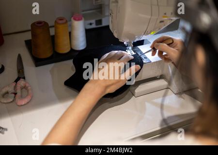 Woman is sewing black beanie hat on a sewing machine, using a sewing machine on the sewing table Stock Photo
