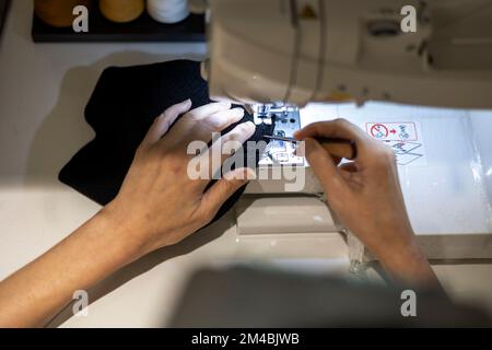 Woman is sewing black beanie hat on a sewing machine, using a sewing machine on the sewing table Stock Photo