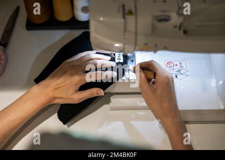 Woman is sewing black beanie hat on a sewing machine, using a sewing machine on the sewing table Stock Photo