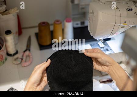 Woman is sewing black beanie hat on a sewing machine, using a sewing machine on the sewing table Stock Photo