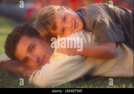 Father and son laying in the grass with son on fathers back, both looking at the camera Stock Photo