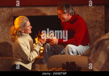 Older couple sitting in front of a fireplace fire sharing a drink and clicking glasses together Stock Photo