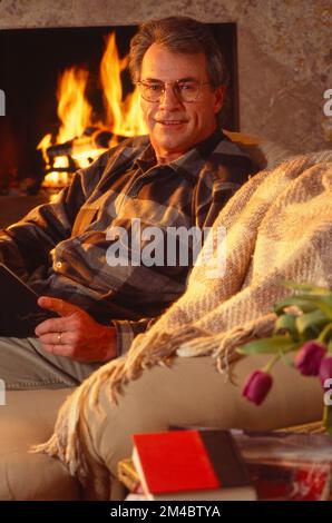 Older man sitting on the couch in front of a fireplace pausing to look up while reading a book Stock Photo