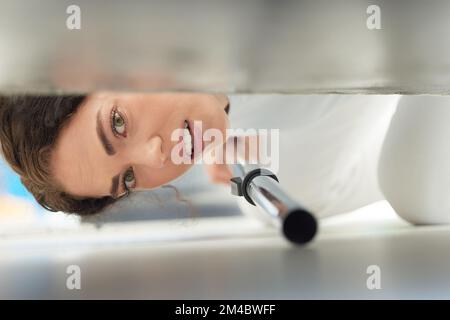 Cleaning, vacuum and face of woman under sofa for housework or hygiene. Spring cleaning, machine and portrait of female cleaner vacuuming floor to Stock Photo