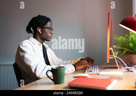 Focused African man freelance web developer writing code, using computer, sitting at home office Stock Photo