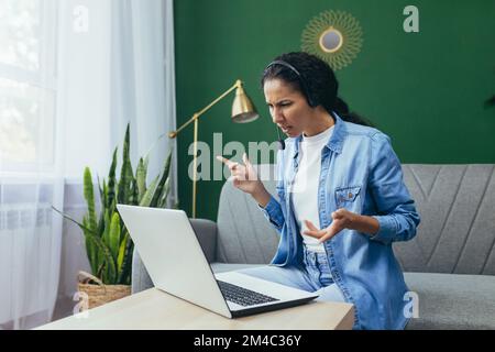 Portrait of frustrated and upset woman at home, Hispanic woman working remotely looking at laptop at work and video call headset sitting on sofa in living room. Stock Photo