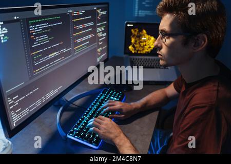 Profile view of young developer programming at home office, man in shirt and glasses writing code on pc computer checking database. Program development and data science concept  Stock Photo