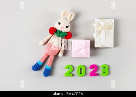 Knitted children's toy bunny or rabbit with pink and beige gift boxes and numbers 2023 on a gray background. The symbol of 2023. Stock Photo