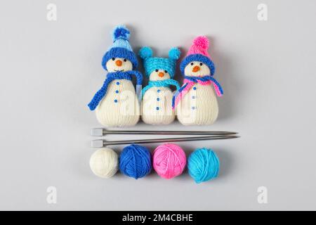 Three knitted snowmen in blue and pink hats with scarves with balls of thread and knitting needles on a gray background. Winter knitting creativity, t Stock Photo