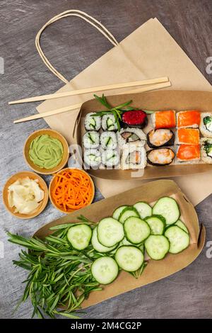 Takeaway food. Rosemary sprigs and sliced cucumbers, sushi in paper plates. Seasonings in bowls. Paper bag. Flat lay. Brown background. Stock Photo