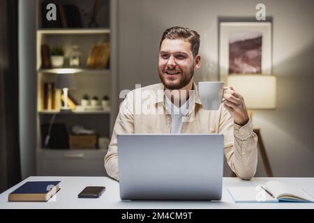 Happy young man holding cup of hot everage and smiling to camera, sitting at desk with laptop. Remote developer finishing project Stock Photo