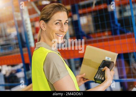 Happy female worker with protective vest and scanner, scans bar-code of package, standing at warehouse of freight forwarding company Stock Photo
