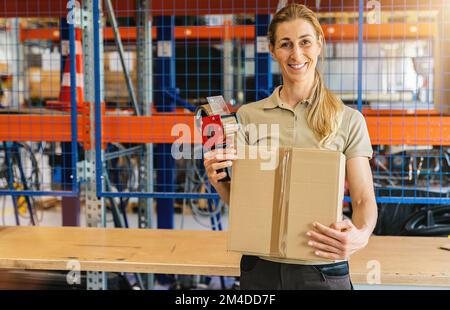 Happy female worker holding packing case after sealing it with adhesive tape at warehouse of freight forwarding company Stock Photo