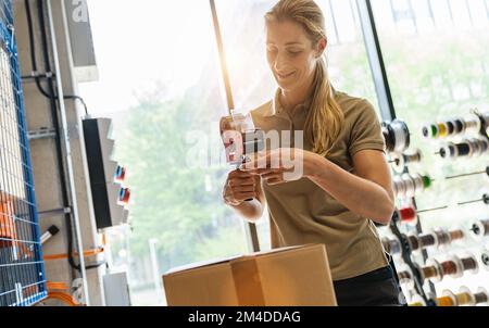 smiling female worker sealing shipping box with tape dispenser and adhesive tape at warehouse of freight forwarding company Stock Photo