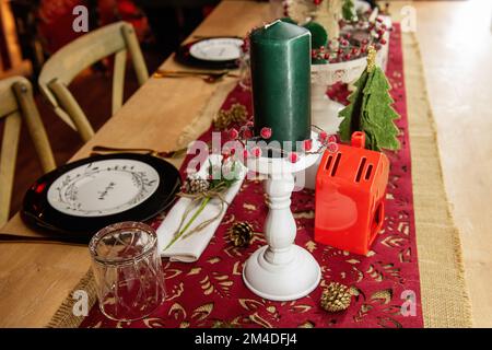 Festive Christmas table setting. On a wooden table lies a red runner, black plates on white says Enjoy, golden forks, spoons, knives. New year green c Stock Photo