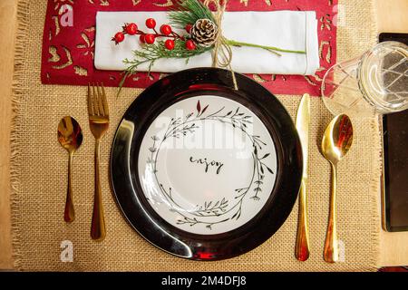 Festive Christmas table setting. On a wooden table lies a red runner, black plates on white says Enjoy, golden forks, spoons, knives. New year green c Stock Photo