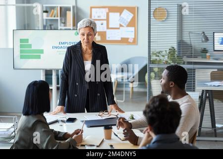Portrait of senior woman heading business meeting in office and standing at table briefing team Stock Photo