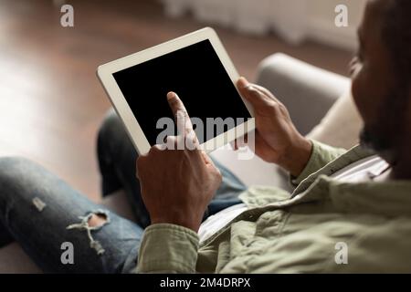 Smiling adult black guy uses tablet with blank screen for online work, business, sits on sofa in living room Stock Photo