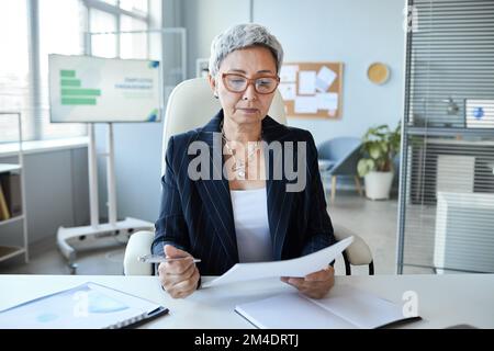 Portrait of senior woman as female boss sitting at workplace in office and reading document Stock Photo