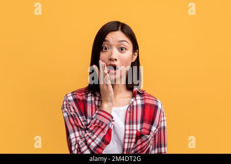 Shocked young asian lady looking at camera with open mouth and touching cheek, emotionally reacting to news Stock Photo