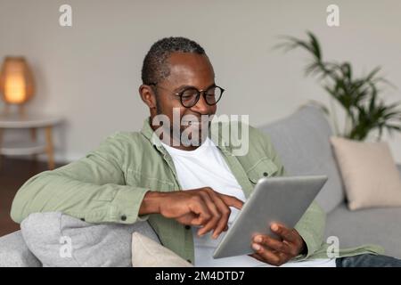 Happy adult black guy in glasses uses tablet for online work and business, sits on sofa in living room interior Stock Photo