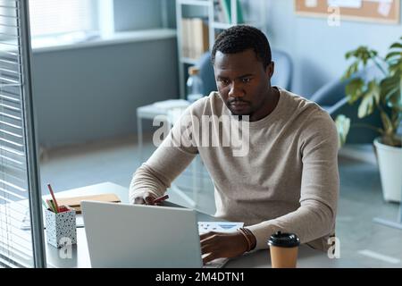 Portrait of focused black man using laptop in office behind glass partition wall, copy space Stock Photo