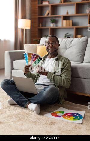 Cheerful adult black man in glasses uses computer, works with color palettes for design, sits on floor Stock Photo