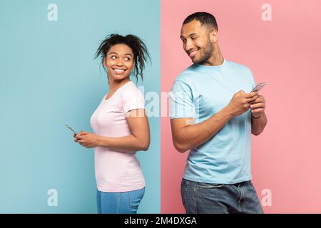 Curious black man using smartphone, looking and peeking at his girlfriend's phone, standing on pink and blue background Stock Photo
