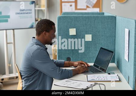 Side view portrait of black man working with laptop at workplace cubicle in office Stock Photo