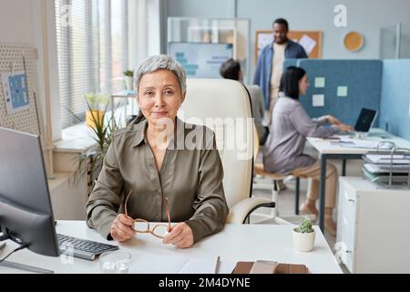 Front view portrait of elegant senior businesswoman working at desk in open office setting and smiling at camera Stock Photo