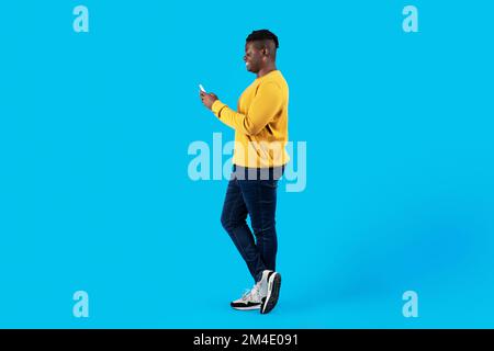 Side View Of Young Black Male With Smartphone Standing Over Blue Background Stock Photo