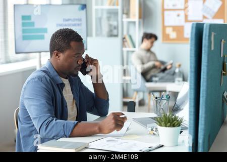 Side view portrait of black man speaking on phone while working at desk in office, copy space Stock Photo