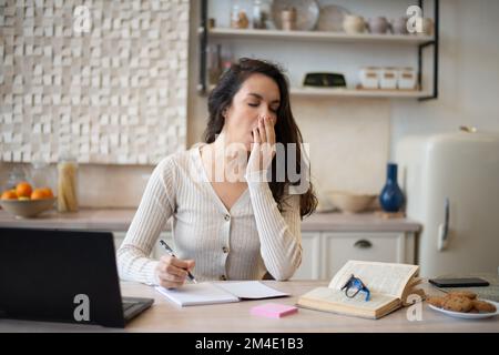 Tired woman working with laptop and yawning, studying online in light kitchen interior, free space Stock Photo