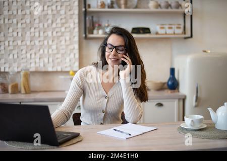 Portrait of happy caucasian woman working from home, talking on cellphone and using laptop computer, sitting in kitchen Stock Photo