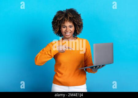 Smiling young black lady shopping online, holding laptop on blue Stock Photo