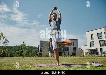 Full length portrait of young man doing yoga outdoors balancing tree position in sunlight, copy space Stock Photo