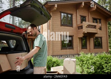 Side view portrait of young man unloading boxes from car trunk while moving into new wooden house in nature, copy space Stock Photo
