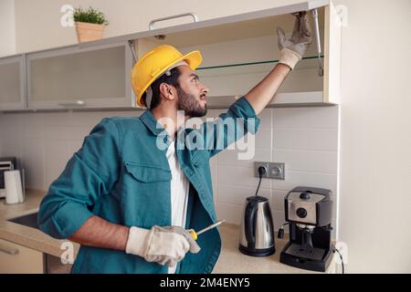 Focused professional worker in uniform is assembling furniture on kitchen. Repair concept Stock Photo