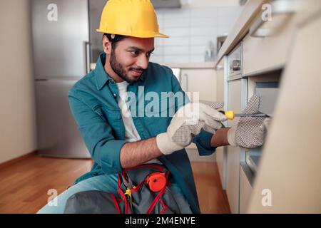 Smiling professional worker in uniform is assembling furniture on kitchen. Repair concept Stock Photo
